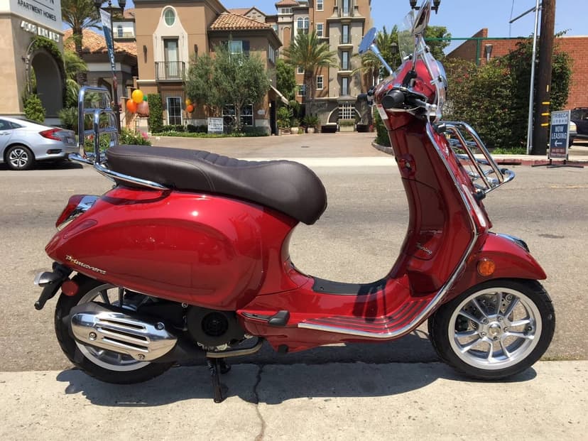 Metallic red Vespa Primavera scooter with chrome racks parked on a sunny urban sidewalk under a clear sky.