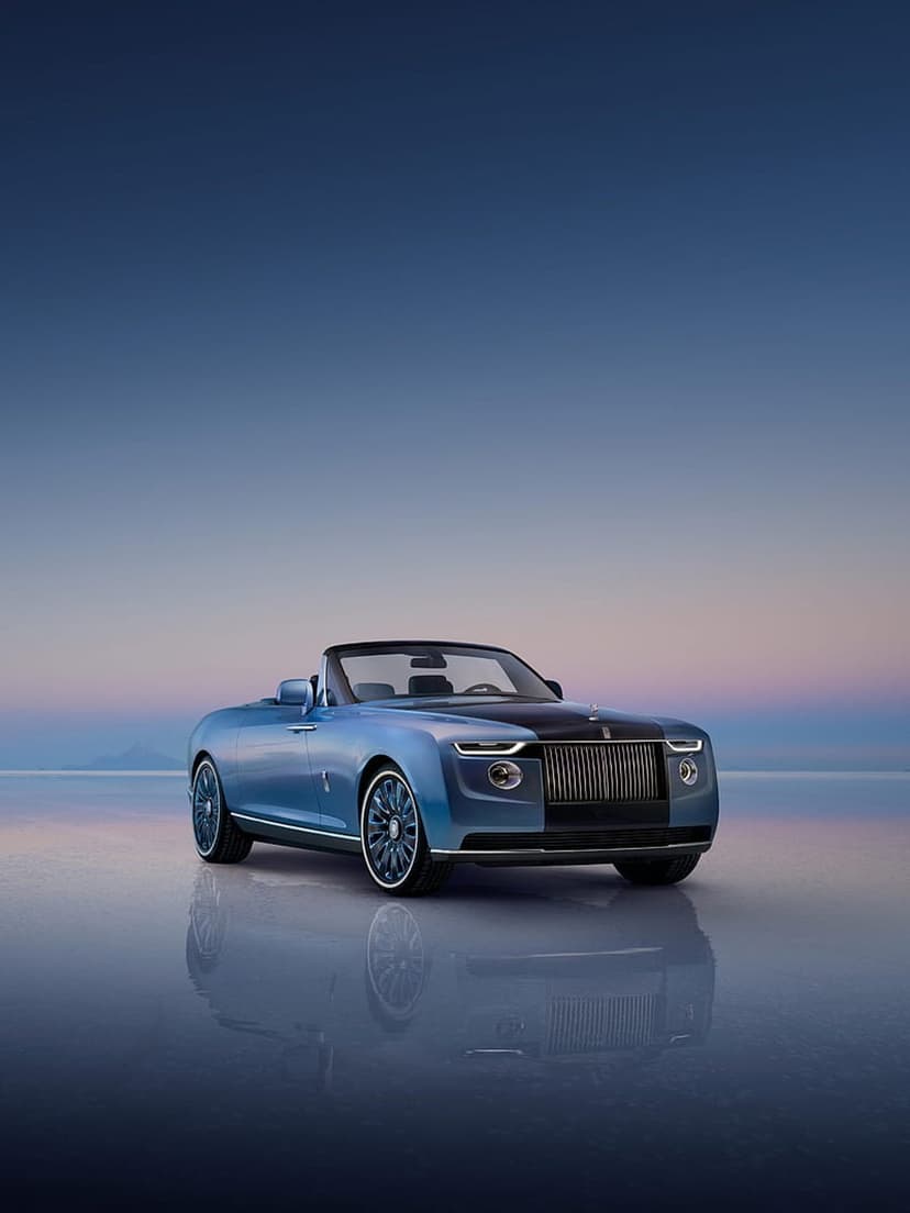 Blue Rolls-Royce Droptail convertible sits on a reflective floor against a purple and blue twilight sky.