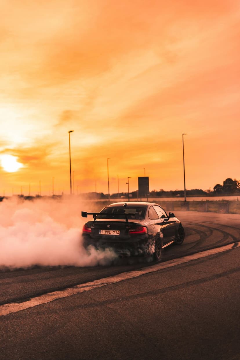 Black sports car slides sideways on a track, emitting thick white smoke under a glowing orange sunset sky.