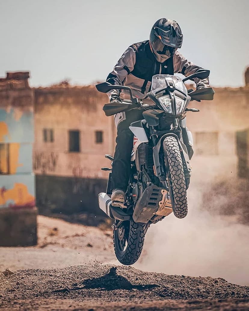 Motocross rider in full gear jumps a dirt bike over a dusty desert mound with old buildings in the background.