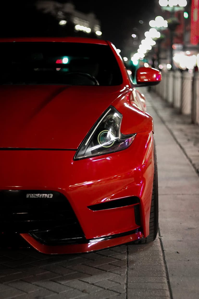 Red Nissan 370Z Nismo sports car parked on a night city street with glowing headlights and bokeh lights.