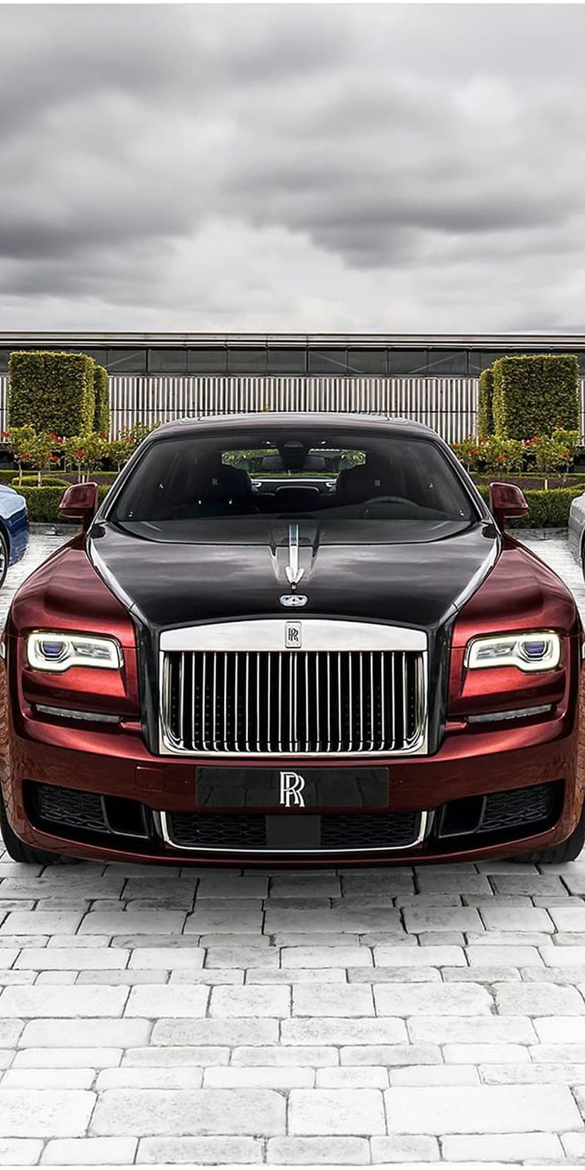 Rolls-Royce Ghost with red paint and a black hood parked on a stone driveway in front of a modern glass building.