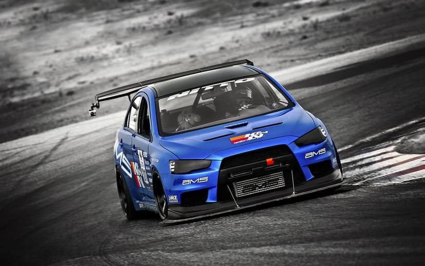 Blue Mitsubishi Evolution race car with a large rear wing cornering on a desaturated asphalt track.