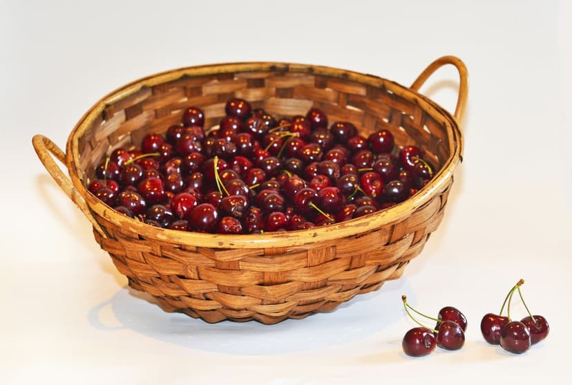 Ripe dark red cherries with green stems spill out of a rustic wicker basket onto a flat, white surface.