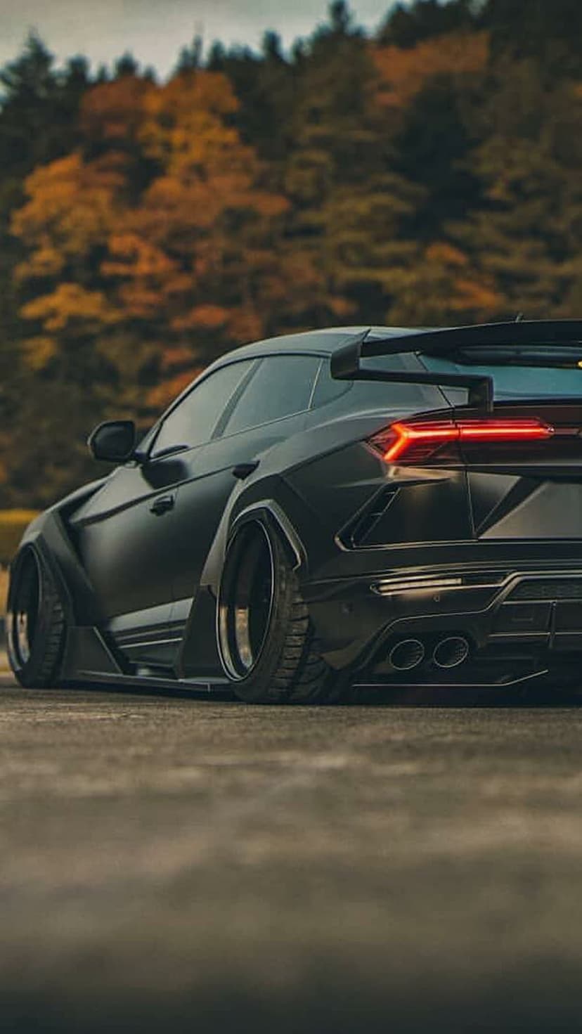 Matte black Lamborghini Urus widebody with glowing red taillights parked against blurred autumn trees.