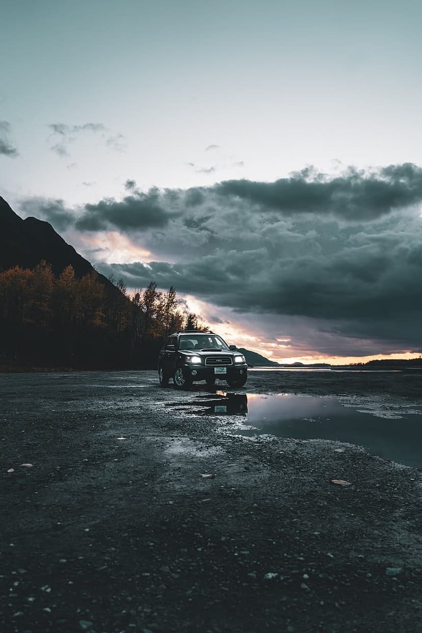Subaru Forester SUV parked on wet gravel by a calm lake under a dramatic stormy sky with orange sunset light.