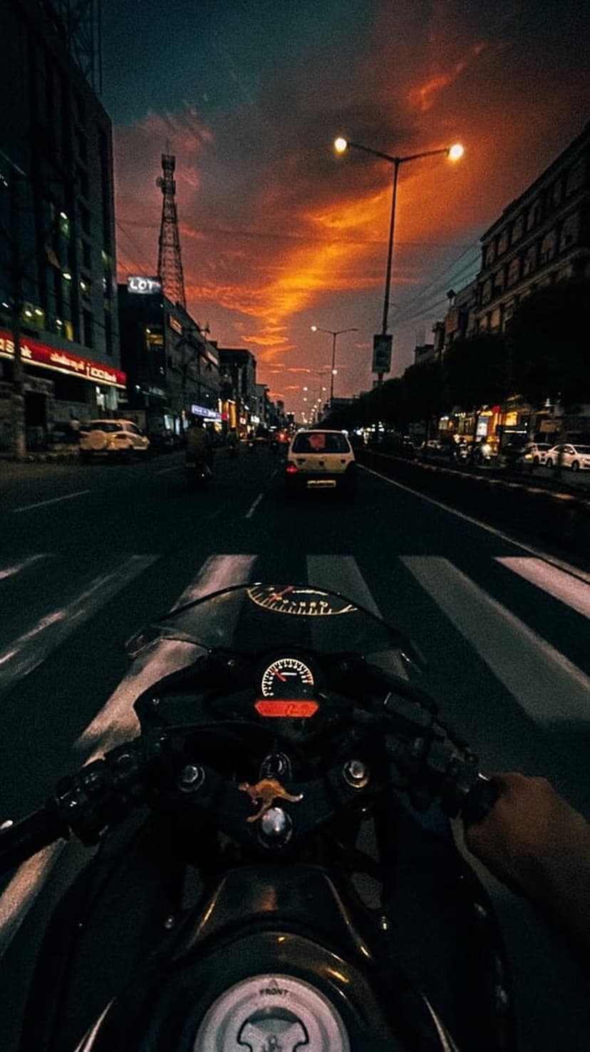 Motorcycle handlebars steer toward a vibrant orange sunset on a dark urban street with glowing traffic lights.