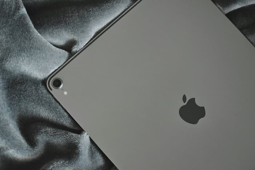 Silver iPad Pro with an Apple logo lies on a textured grey blanket, showing the camera lens and metallic finish.