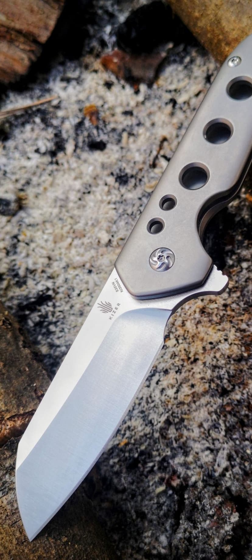 Kizer folding knife with a titanium handle and circular cutouts sits on a bed of grey ash and charred wood pieces.
