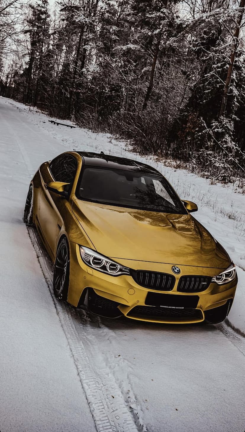 Gold BMW M4 coupe navigates a winding snow-covered road flanked by frost-dusted evergreen trees in winter.