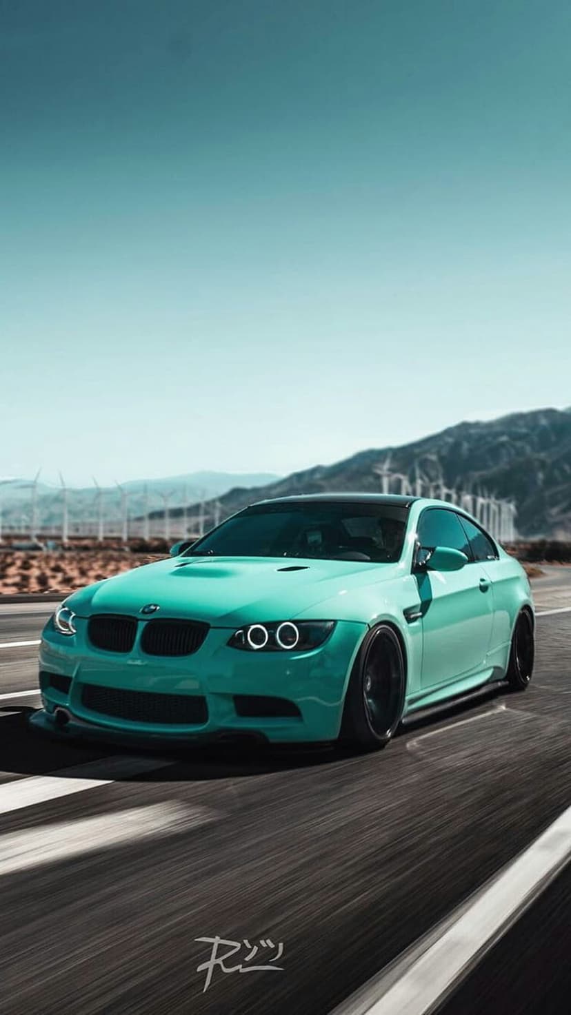 Teal BMW M3 coupe with black rims driving on a desert road featuring wind turbines and distant mountain peaks.