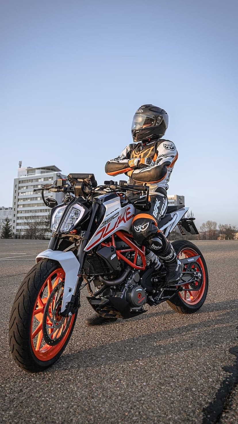 Motorcyclist in a full racing suit sits on a KTM Duke 390 with orange rims under a clear blue sky.