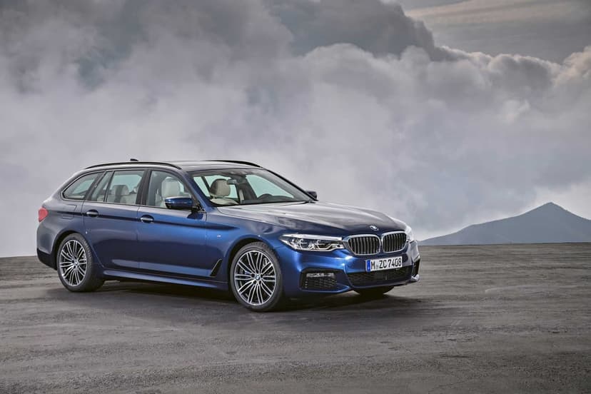Blue BMW 5 Series Touring parked on dark textured ground against a backdrop of heavy grey and white clouds.