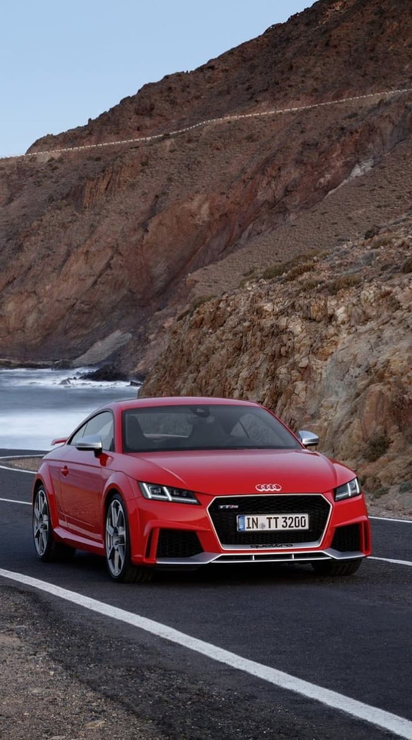 Red Audi TT RS coupe drives on a winding coastal road beside a rocky cliff and blue ocean at a low angle view.