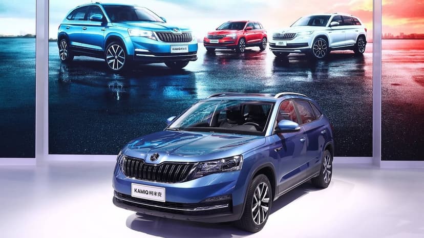 Front view of a blue Skoda Kamiq SUV in a showroom with a sunset background screen showing other car models.