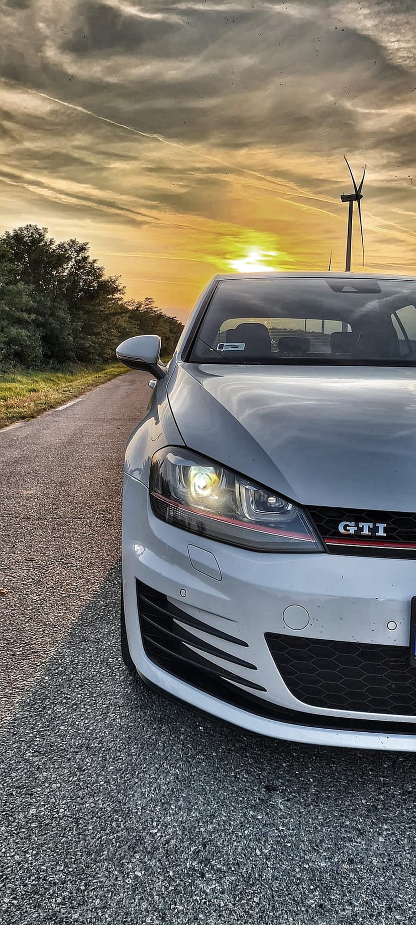 Silver Volkswagen Golf GTI on a rural road at sunset with wind turbines and glowing headlights in the background.