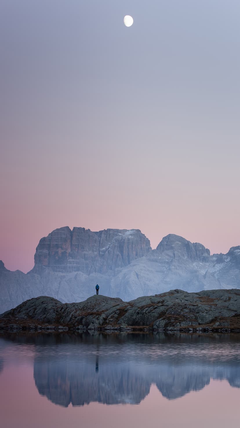 Lone figure standing on a rocky shore reflects in a mountain lake under a glowing moon and pink twilight sky.