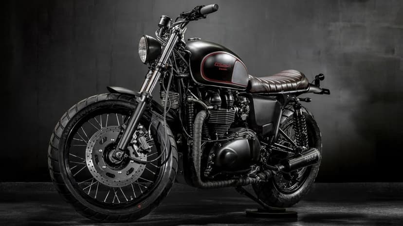 Custom matte black Triumph motorcycle with a brown leather seat and wrapped exhausts in a dark studio setting.