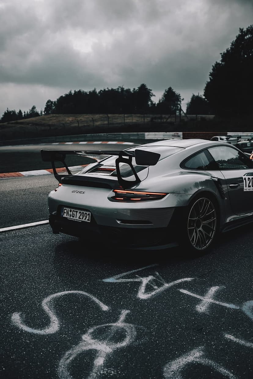 Silver Porsche 911 GT3 RS with glowing red taillights on a wet race track under a dark, moody and cloudy sky.