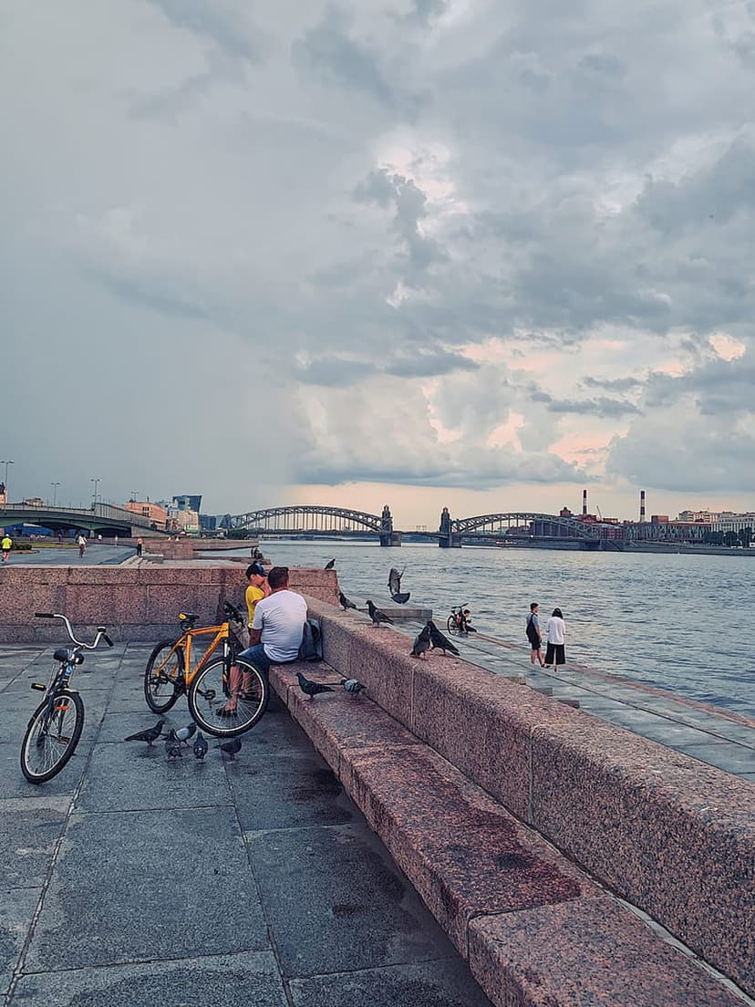 People resting on stone benches near a yellow bicycle along a cloudy riverfront with a large bridge behind them.
