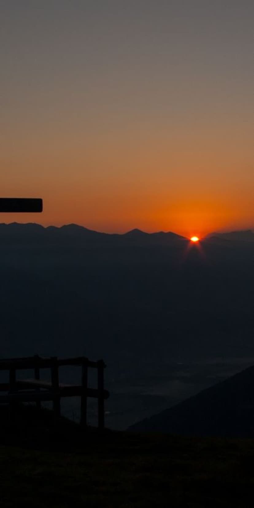 Silhouetted mountain peaks rise above a cloud-filled valley as a bright orange sun emerges during a vivid dawn.