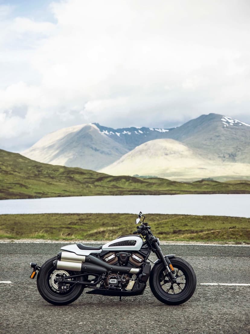 White Harley-Davidson Sportster S parked on a road with misty mountains, a lake, and green hills in the distance.