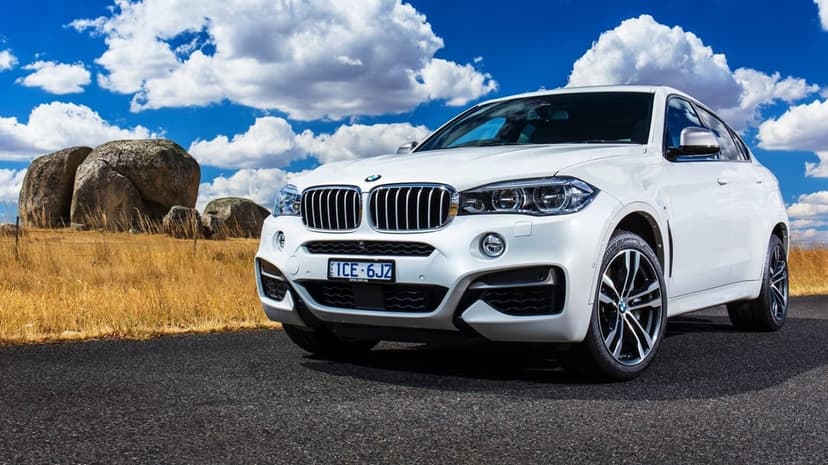 White BMW X6 SUV parked on an asphalt road with large rocks, dry grass, and a cloudy blue sky background.