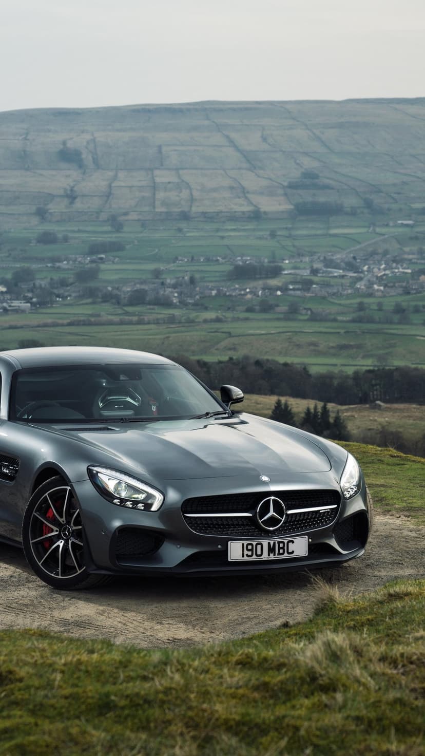 Grey Mercedes-AMG GT S sports car parked on a grassy hill overlooking a vast rural valley landscape.