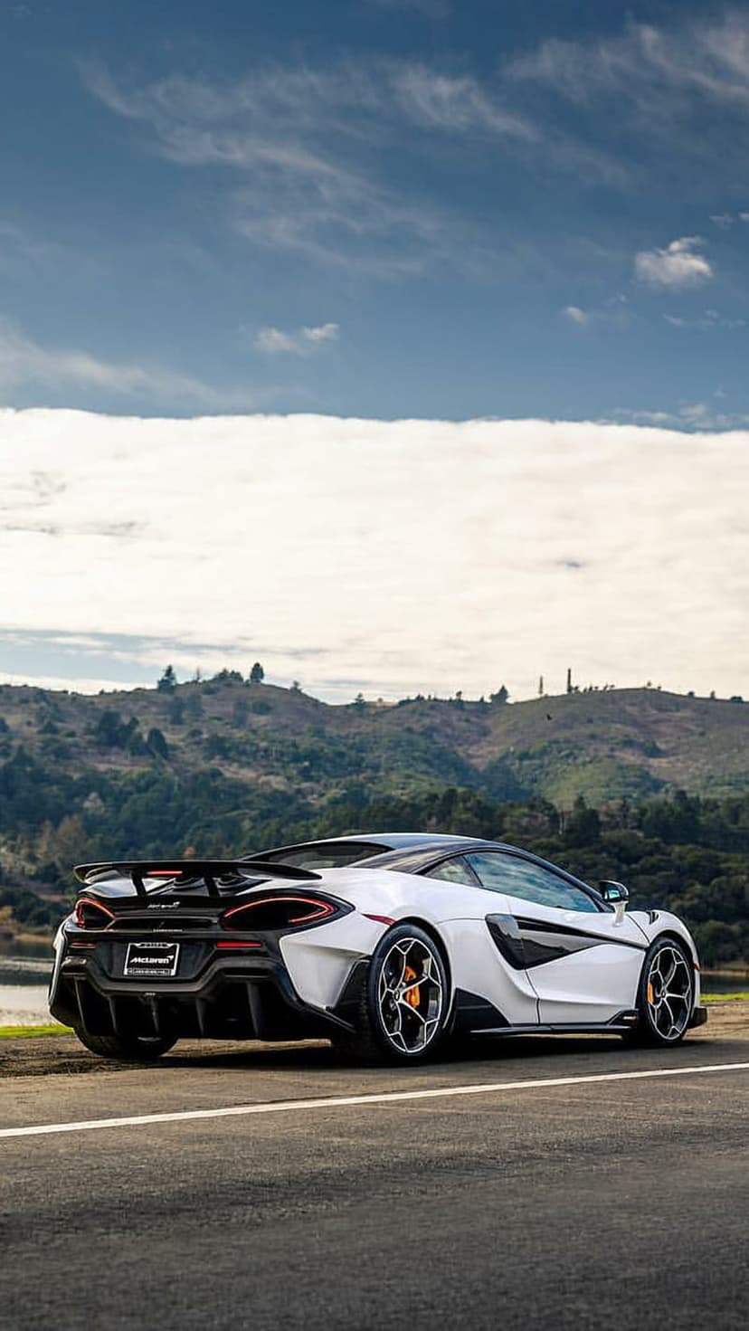 White McLaren 600LT Spider rear view with a large wing and orange calipers on a road through green hills.