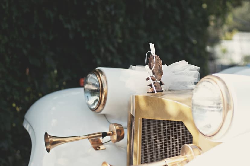 Classic white car front with a gold grille, brass trumpet horn, and an eagle figurine draped in white ribbons.