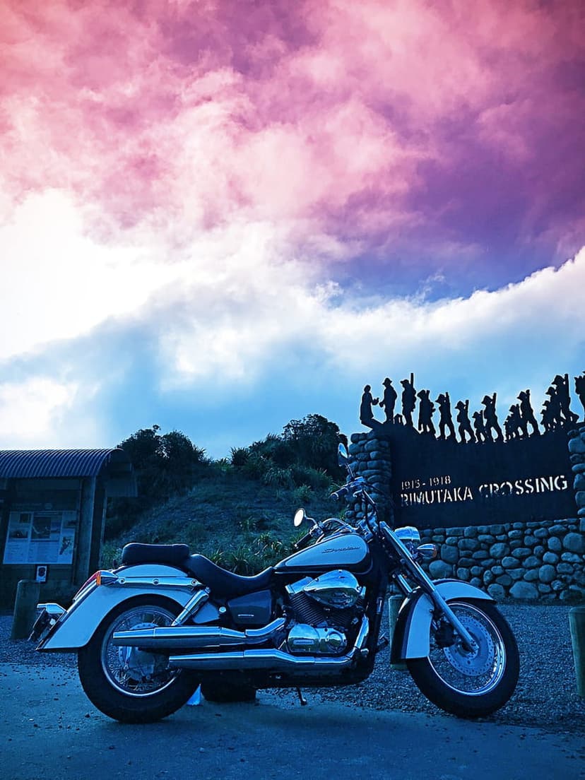 Chrome motorcycle parked by the Rimutaka Crossing memorial under a purple and pink sunset sky with silhouettes.