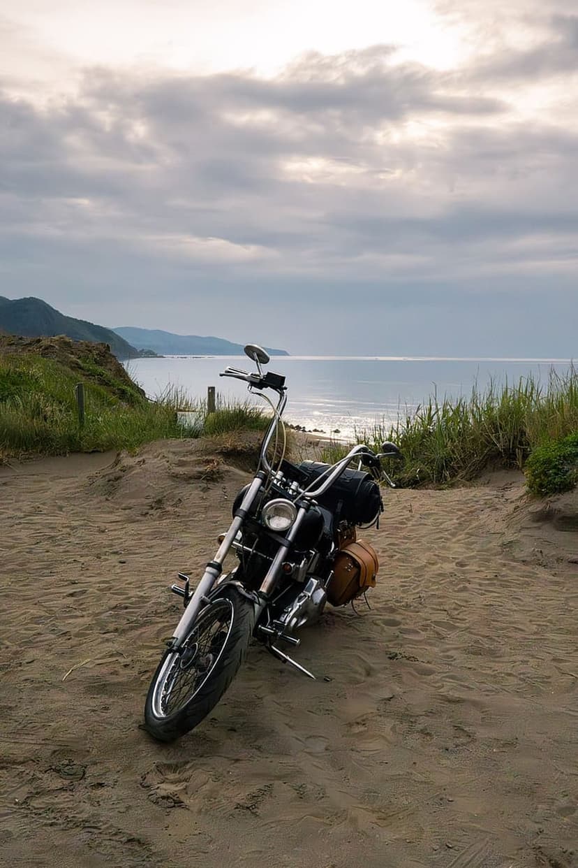 Black vintage motorcycle with chrome accents parked on sandy dunes beside beach grass and a calm coastal ocean.