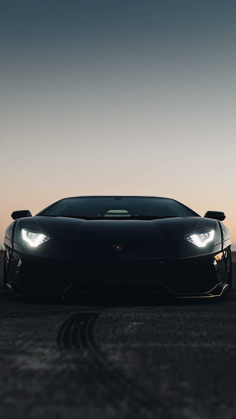Black Lamborghini Aventador with glowing LED headlights on dark asphalt under a moody twilight sky.