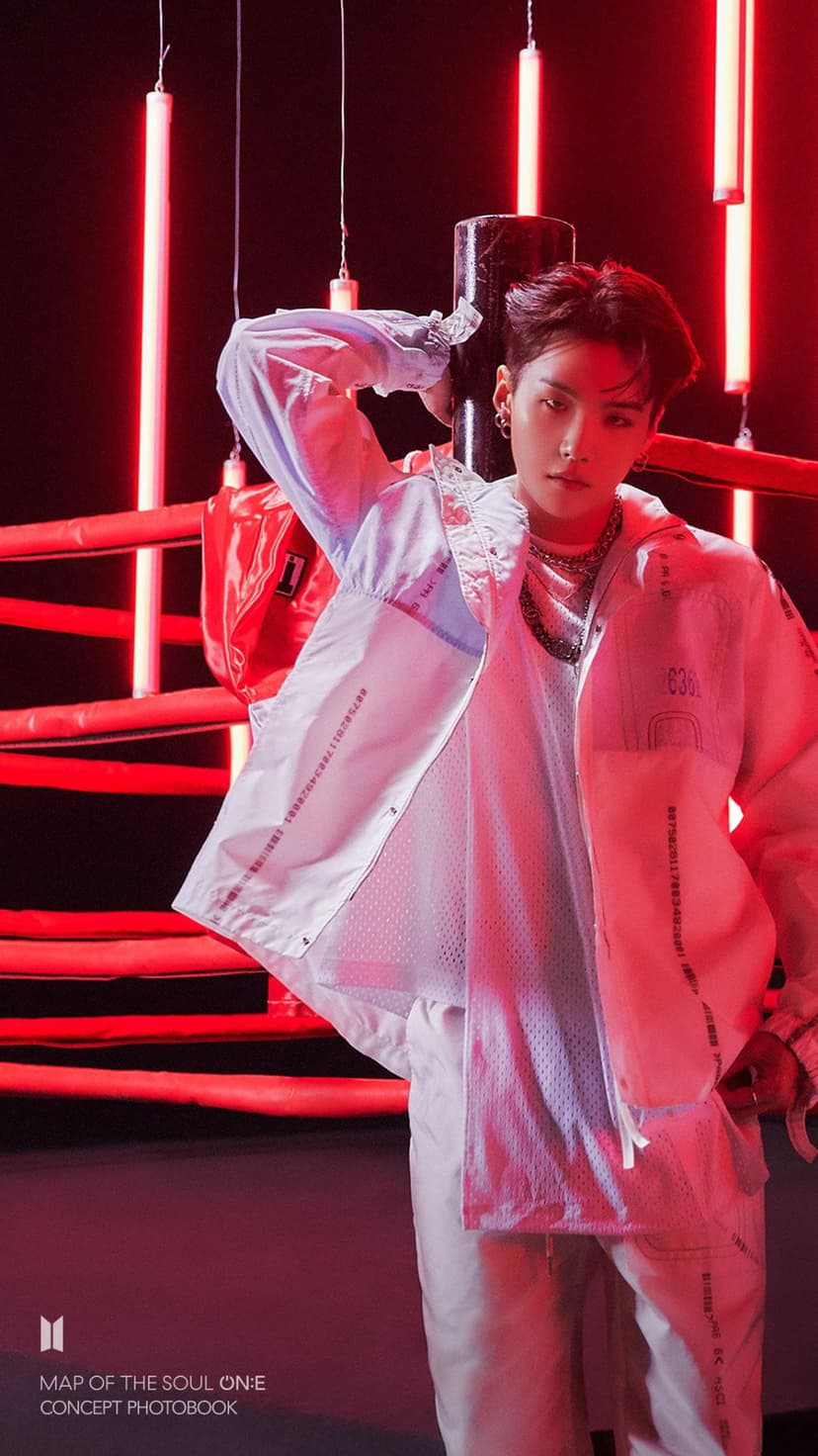 J-Hope of BTS in a light jacket and mesh top stands inside a boxing ring illuminated by red neon lights.