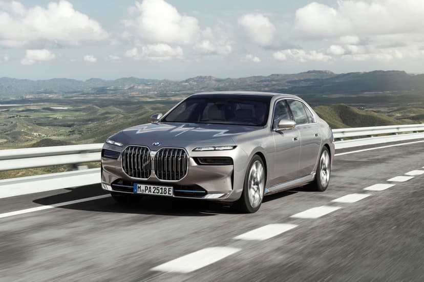 Silver BMW i7 luxury electric sedan driving on a winding mountain road under a cloudy blue sky with motion blur.