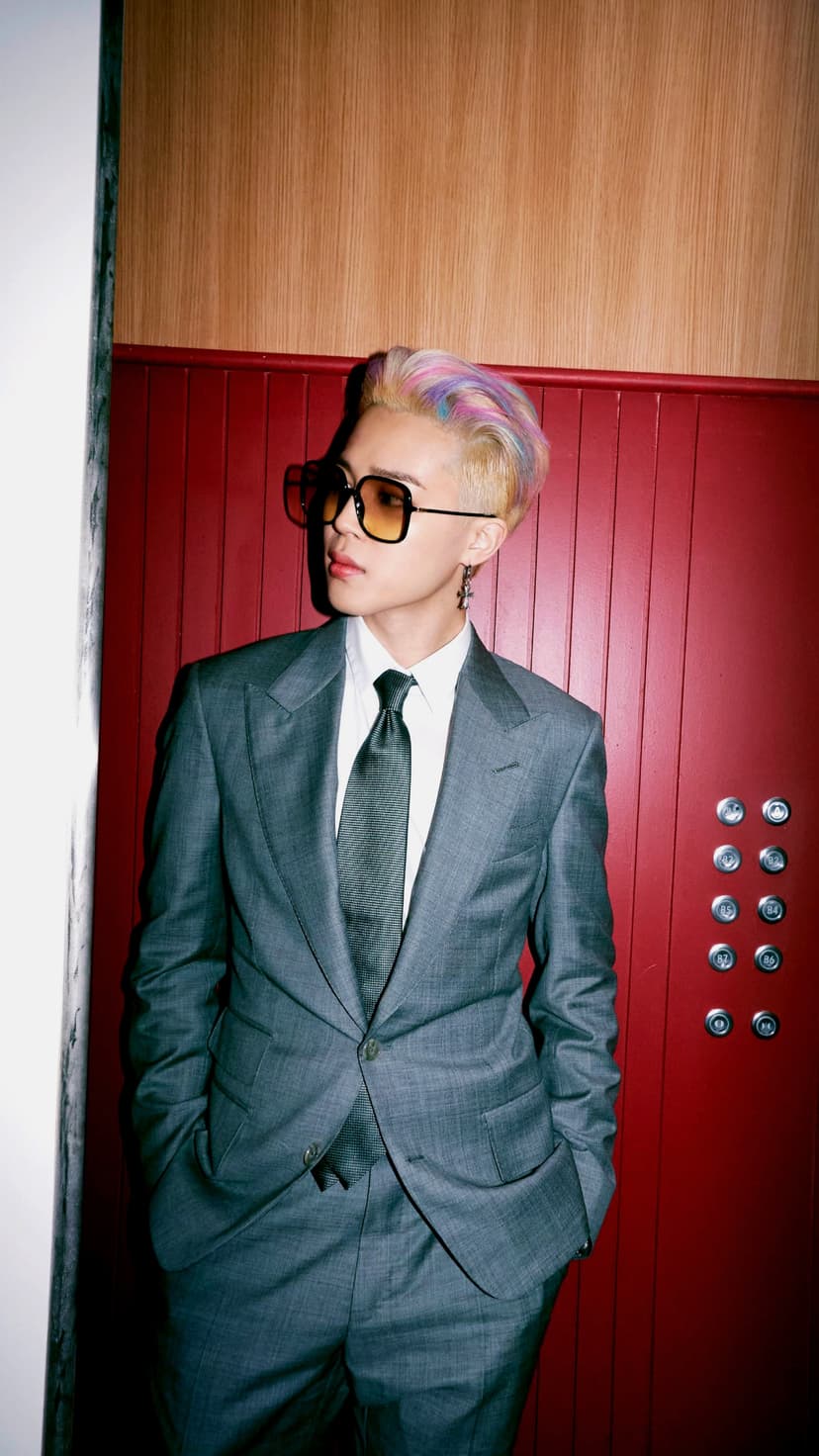 Stylish man in a gray suit with vibrant rainbow hair and yellow sunglasses poses by a red paneled elevator wall.