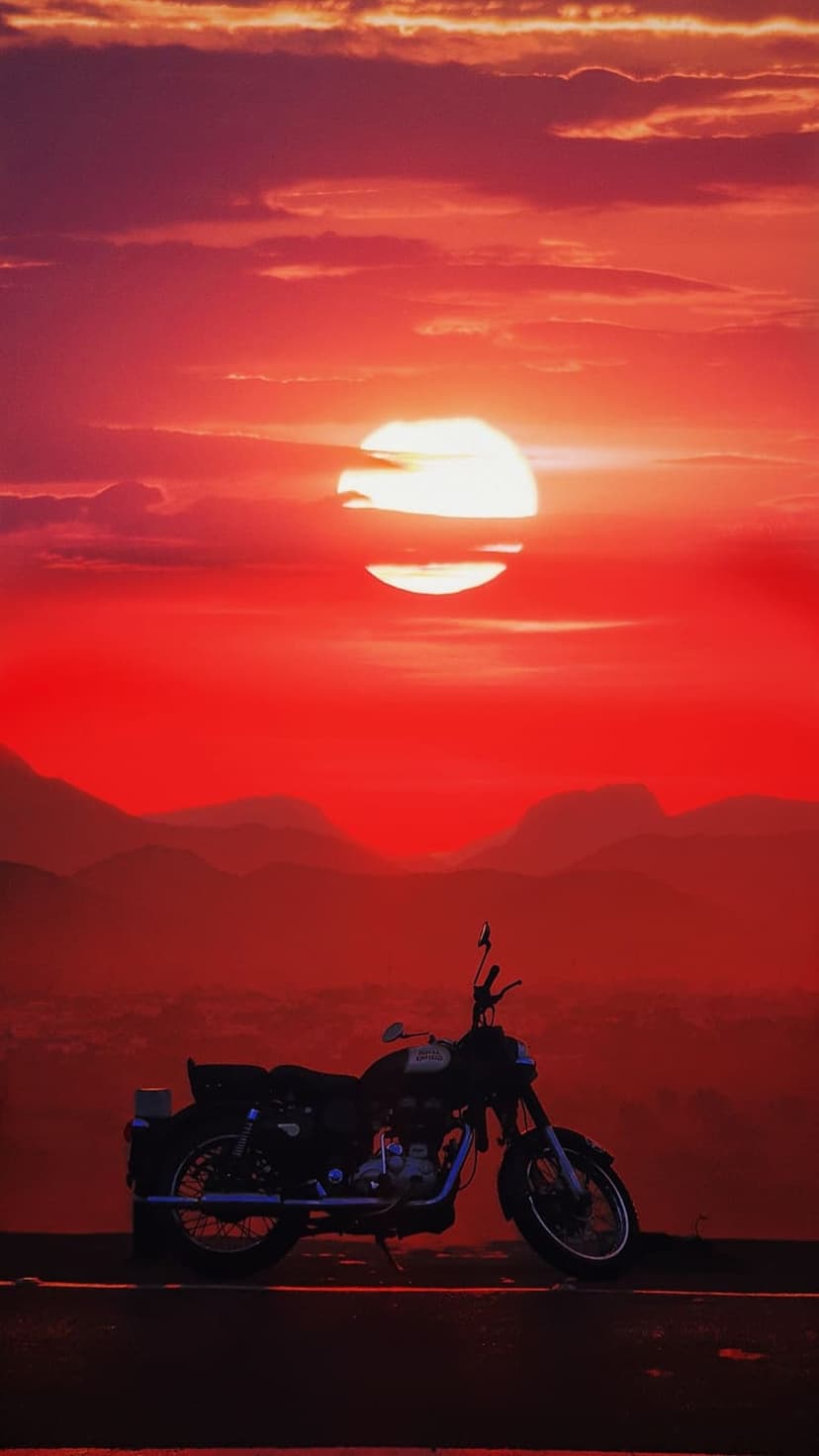 Vintage motorcycle silhouette on a mountain ridge during a deep red sunset with layered clouds and hazy peaks.