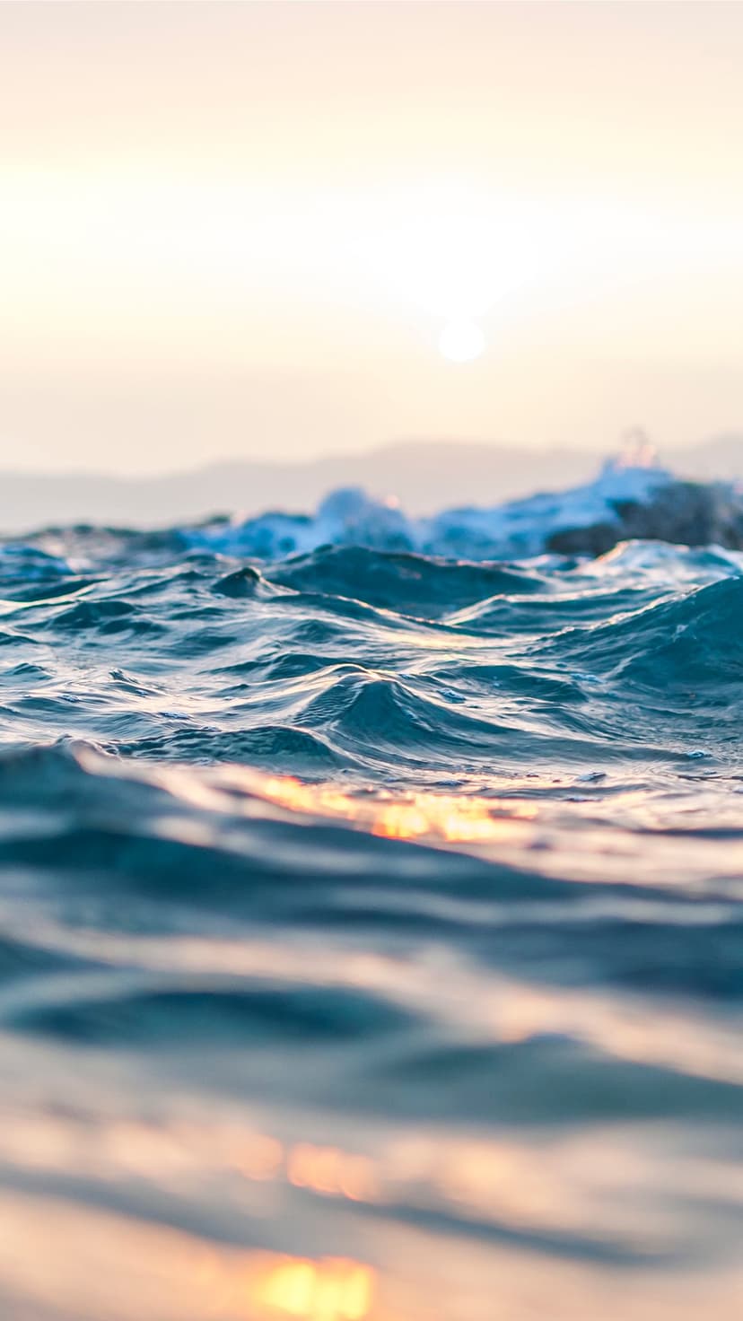 Gentle sea waves reflect orange sunlight beneath distant hazy mountains during a calm evening sunset.