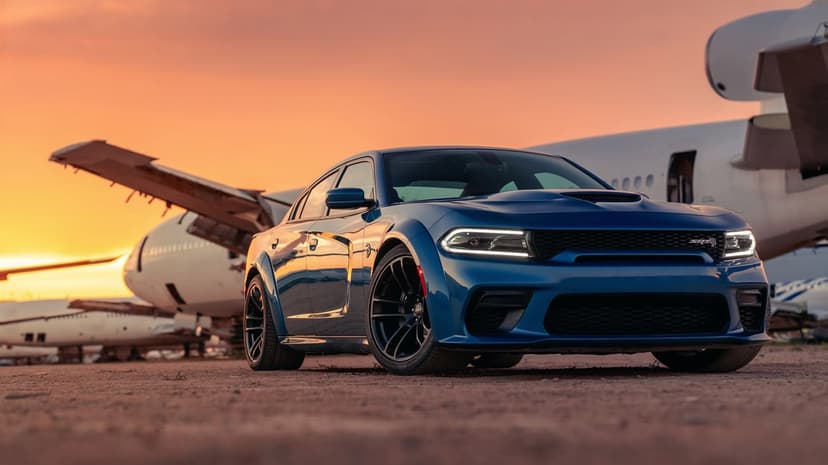 Blue Dodge Charger SRT Hellcat Widebody parked near commercial airplanes during a vibrant orange sunset.