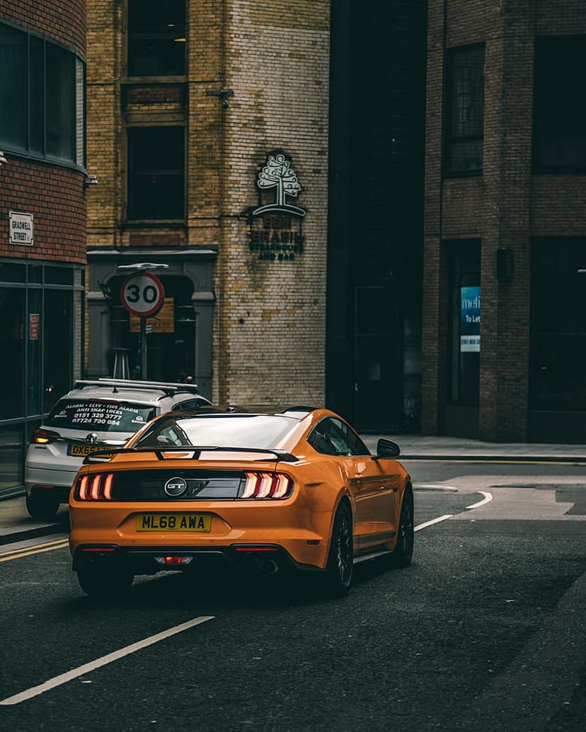 Orange Ford Mustang driving on a city road past a brick building and a 30 mph speed sign in vertical view.