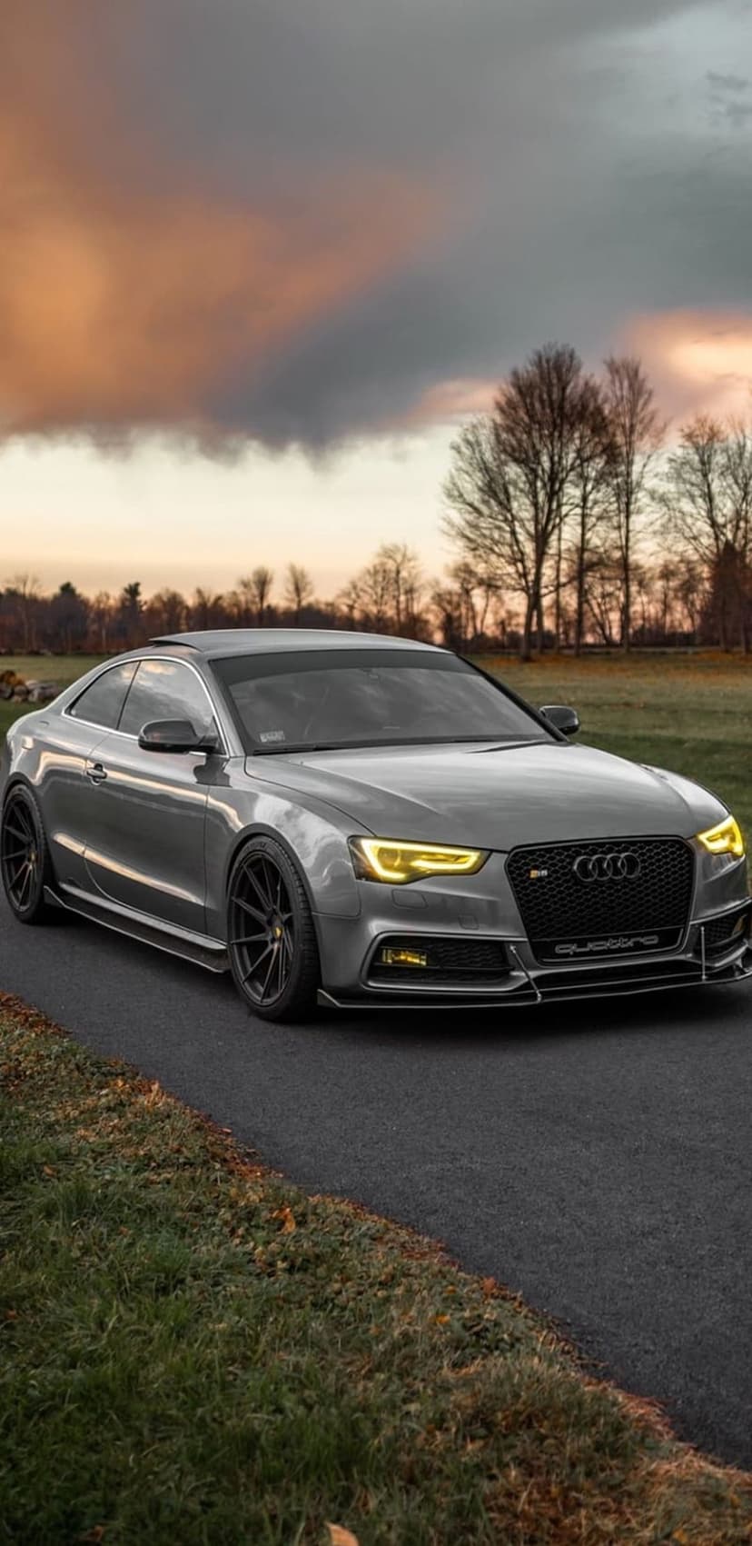 Audi RS5 Coupe with glowing yellow headlights on a winding road beneath a dark, dramatic sunset sky.
