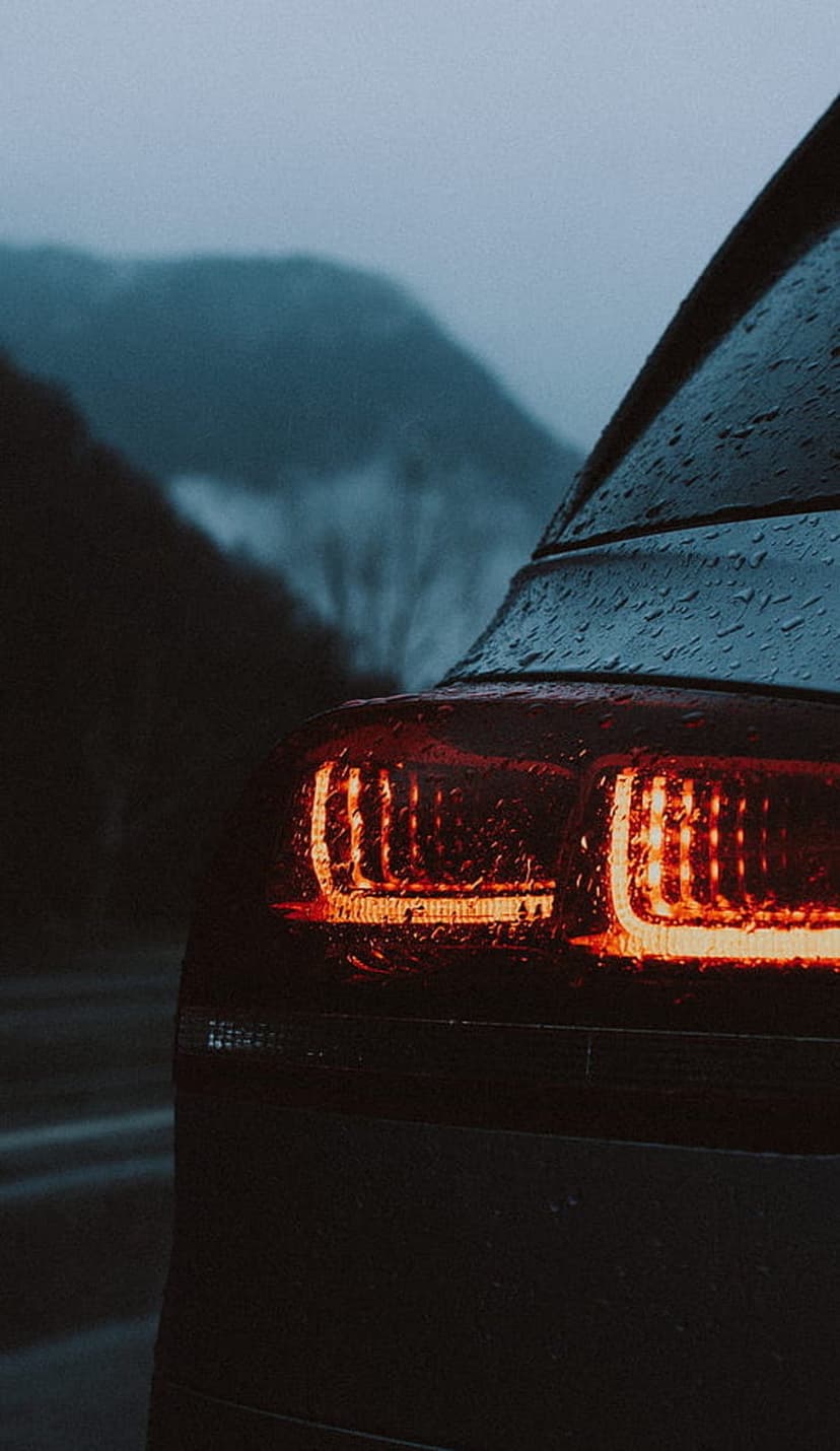 Red car taillight with water droplets glowing against a backdrop of foggy mountains on a rainy evening.