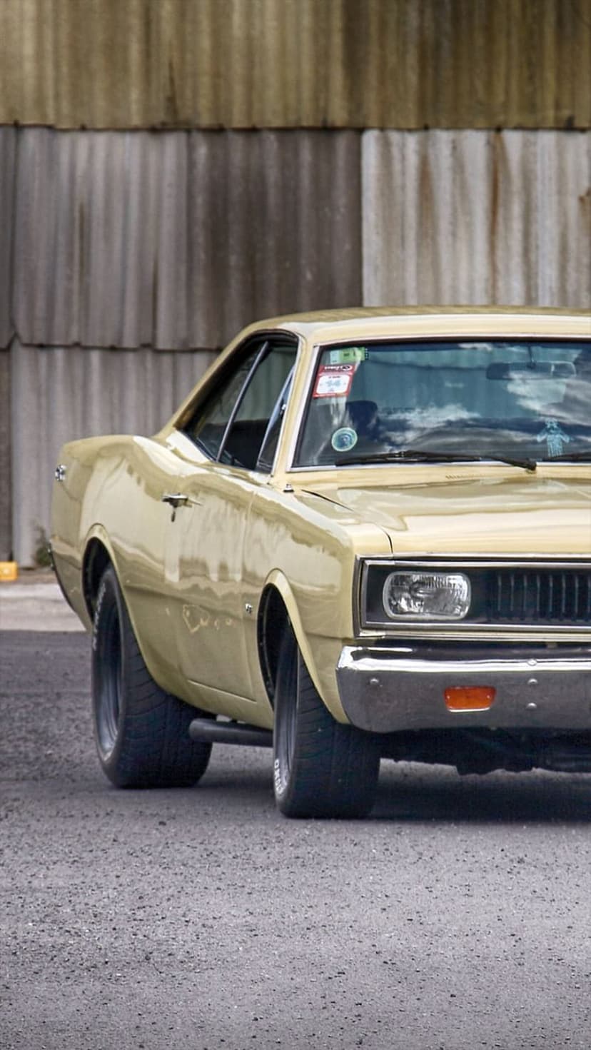 Vintage yellow Dodge Dart with chrome bumpers parked on a gravel path against a rustic wooden barn wall.