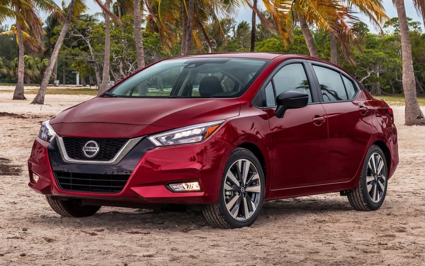 Red Nissan Versa sedan parked on a sandy beach with tropical palm trees and bright daylight in the background.