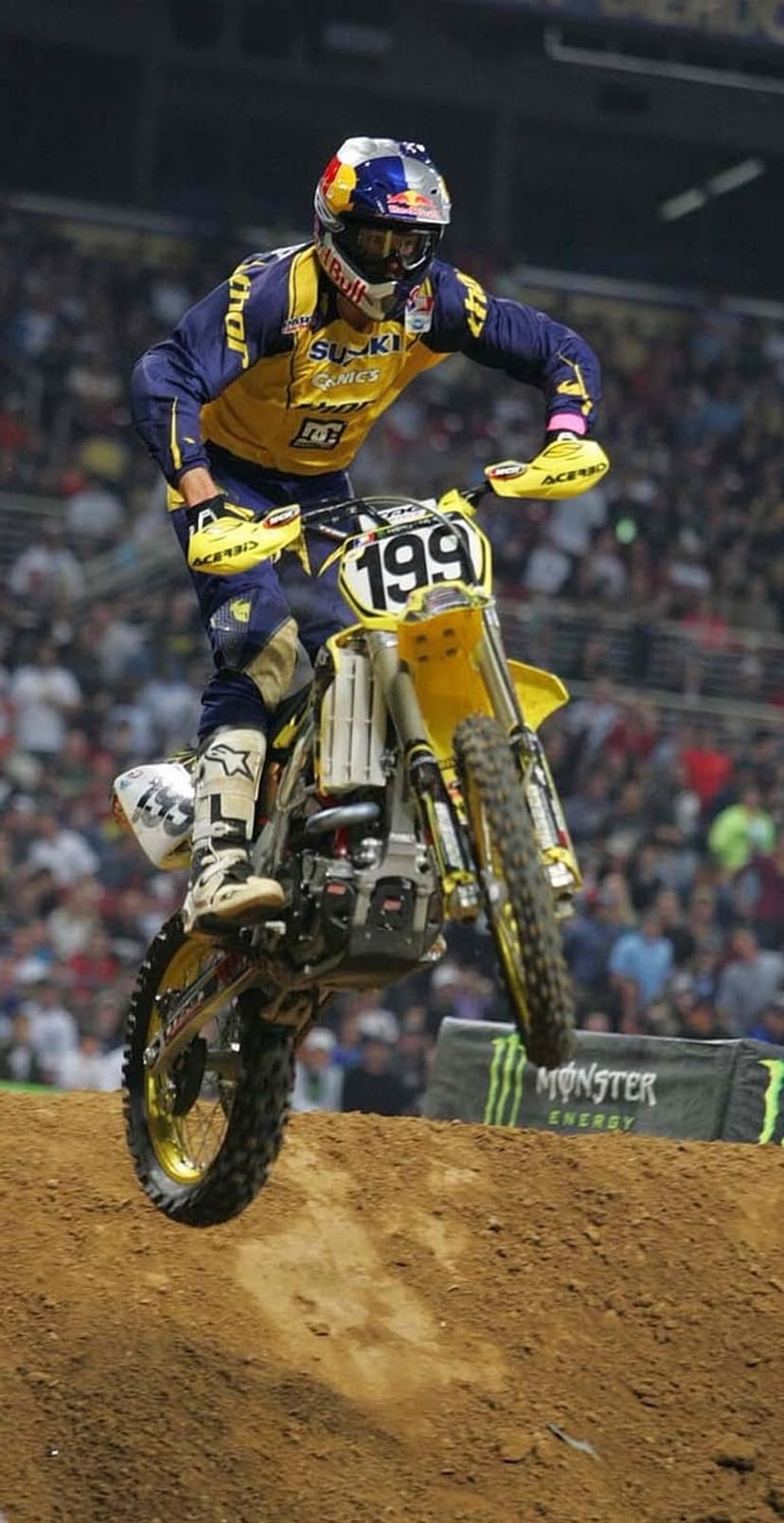 Motocross rider on a yellow Suzuki bike performs a mid-air jump in an indoor stadium during an extreme sports event.