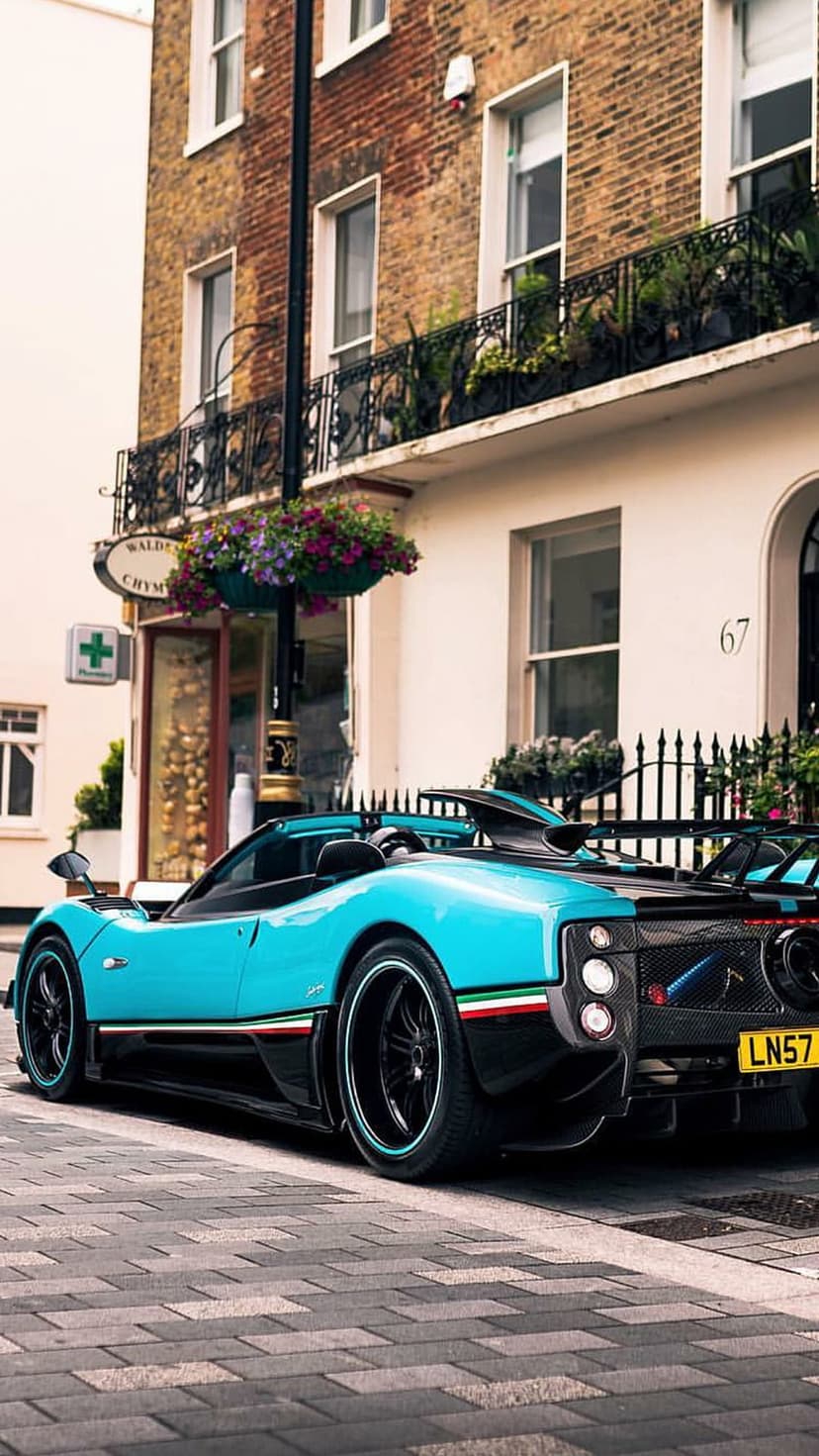 Turquoise Pagani Zonda supercar with Italian stripes on a cobblestone street beside a brick building.