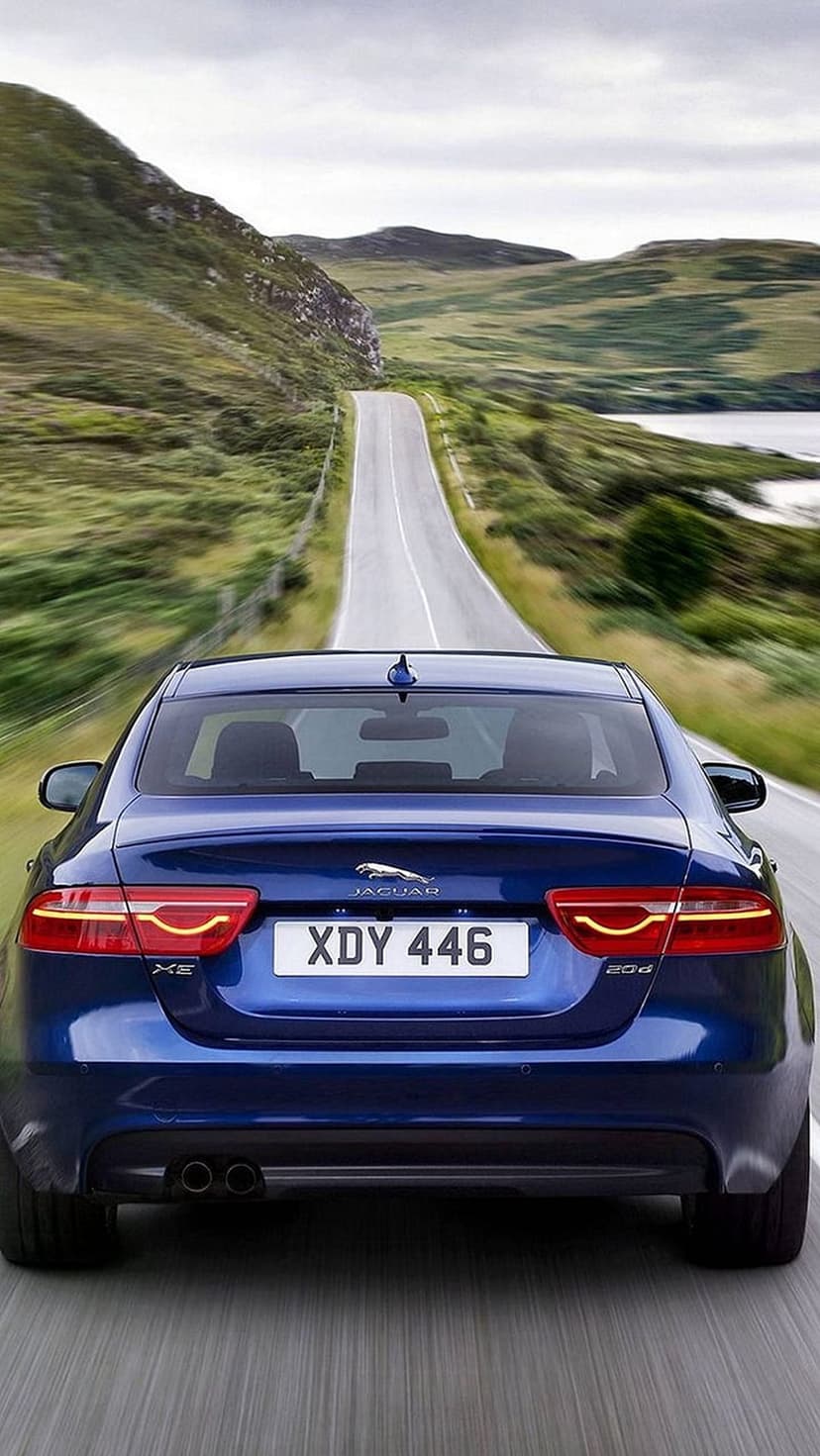 Blue Jaguar XE sedan driving on a winding road through green Scottish hills and a loch under a cloudy sky.