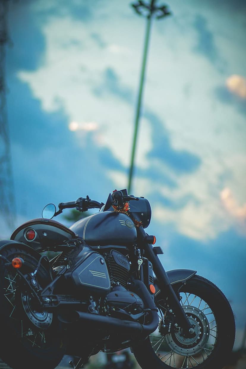 Dark classic motorcycle with chrome accents parked under a dramatic sky featuring pink clouds and a tall lamppost.