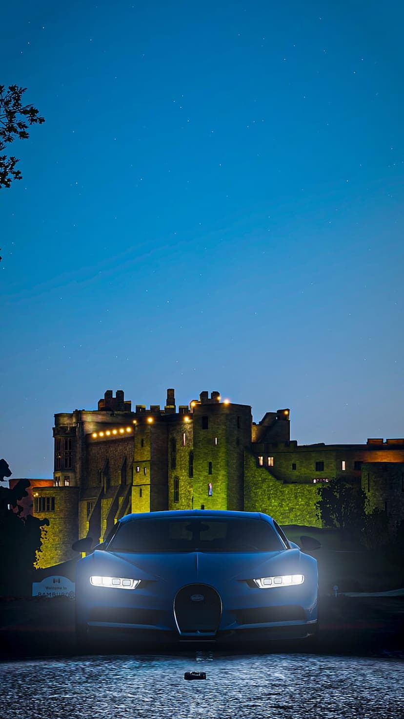 Blue Bugatti Chiron with glowing headlights parked on cobblestones in front of an illuminated stone castle.