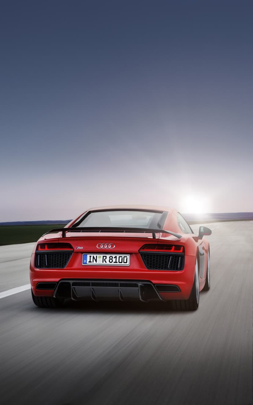Red Audi R8 sports car driving on an open highway toward a vibrant golden sunset with a distant mountain range.
